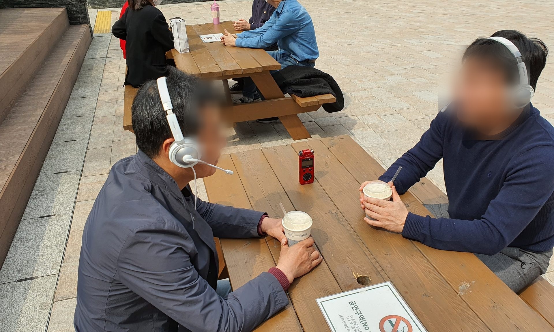야외 테이블에서 헤드셋을 쓰고 소음 환경 대화를 녹음하는 두 참여자