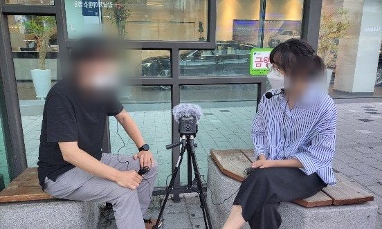 건물 앞 야외에서 마이크 삼각대를 두고 대화를 녹음하는 두 참여자